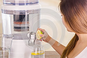 Beautiful woman holding a cut lemon in her hand with a filter system of water purifier on a kitchen background