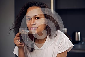 Beautiful woman holding glass cup of freshly brewed americano, smiling looking at camera. Curly haired woman in white