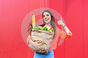 Beautiful woman holding grocery shopping bags on red background showing thumbs up. Happy pretty girl holding bag with groceries