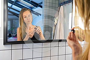 Beautiful woman is holding a jar of cream applying face skin moisturizer, standing in front of the bathroom mirror