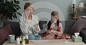 Beautiful woman holding pills blister and talking on smartphone while sitting on sofa with ill son. Young mom having