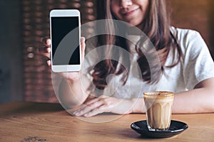 A beautiful woman holding and showing white mobile phone with blank black desktop screen with coffee cup on wooden table
