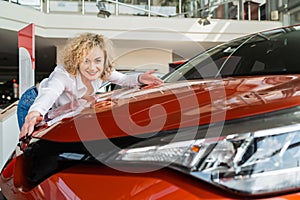 Beautiful woman hugging her new car in dealership. Happy girl rejoices buying a new car.