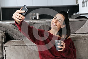 Beautiful woman indoors in home near sofa take a selfie by mobile phone drinking coffee