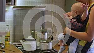 Beautiful woman with an infant boy sitting in a baby carrier cooking soup for a dinner at the kitchen. Mother household