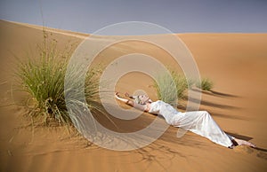Beautiful woman lays in whte dress between tuffets in sand desert