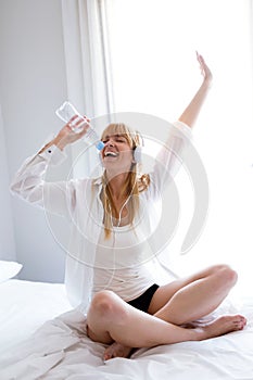 Beautiful woman listening to music and singing with water bottle on the bed at home.