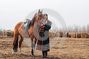 A beautiful woman with long and black hair in a historical hussar costume stands in a field with a horse