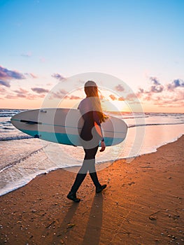 Beautiful woman with long hair go to surfing. Surfgirl with surfboard on a beach at sunset or sunrise. Surfer and ocean