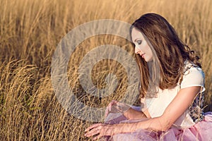 Beautiful woman with long hair wearing white silk top and pink veiling skirt sitting in the yellow field and touching dried grass