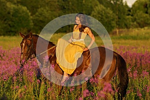 Beautiful woman with long hair in yellow dress riding bareback a brown horse in among purple flowers in green field