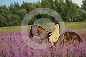 Beautiful woman with long hair in yellow dress riding bareback a brown horse in among purple flowers in green field