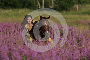 Beautiful woman with long hair in yellow dress riding bareback a brown horse in among purple flowers in green field