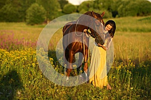 Beautiful woman with long hair in yellow dress standing near brown horse in among purple flowers in green field