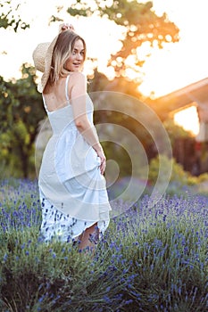 Beautiful Woman in Long White Dress and Hat Stands in Lavender Meadow at Golden Hour. Summer concept