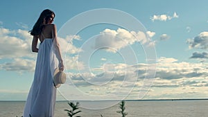 Beautiful woman in long white dress, holding hat in hand, on  background of  sea