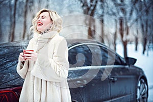 Beautiful woman in luxurious white fur coat drinking hot coffee on snowy winter day and laughing