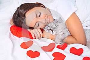 Beautiful woman lying on the bed with red hearts and hugging her lovely cat. Love for pets.