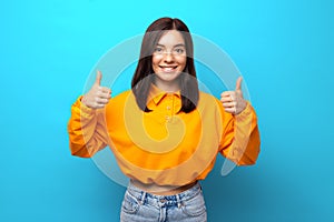 Beautiful woman of mixed ethnicity shows likes with thumbs in orange shirt against blue background