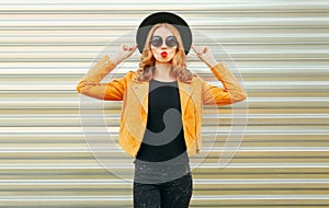 Beautiful woman model blowing red lips sending sweet air kiss wearing yellow jacket, black round hat on wall