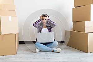 Beautiful woman moving in her new house and unpacking, she is sitting on the floor surrounded by boxes, using a laptop looking sho