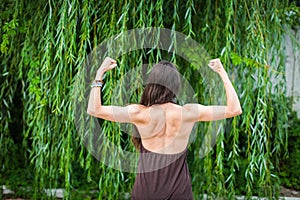 Beautiful woman with naked back over green weeping willow background. Sport girl shows back muscles.