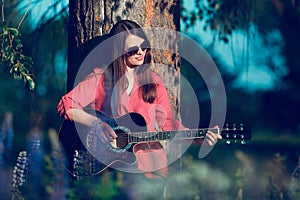 Beautiful woman near the tree and the field with lupine playing guitar