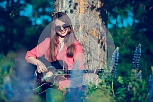 Beautiful woman near the tree and the field with lupine playing guitar