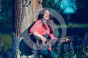 Beautiful woman near the tree and the field with lupine playing guitar