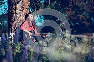 Beautiful woman near the tree and the field with lupine playing guitar