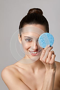 Beautiful woman with nude makeup and naked shoulders posing at grey background with cleaning sponge,