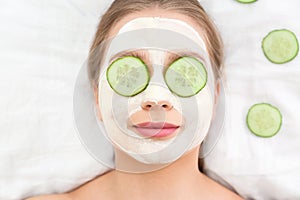 Beautiful woman with organic facial mask and cucumber slices on white fabric