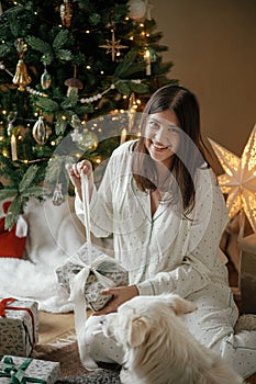 Beautiful woman in pajamas opening stylish christmas gift with cute white dog at decorated tree and fireplace, enjoying cozy