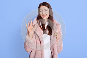 Beautiful woman in pale pink fur coat showing sign ok, keeps one closed, showing her approval, standing against blue wall, wearing