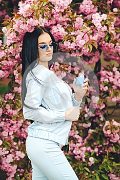 Beautiful woman with perfume bottle in pink flowers. Spring day. Fashionable girl in trendy glasses. Women perfume, fragrance