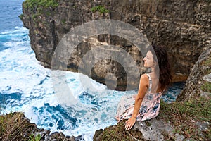 A beautiful woman in a pink dress sits on a cliff above the ocean on the island of Nusa Penida