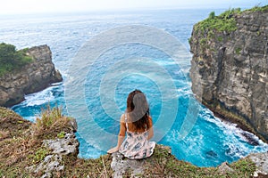 A beautiful woman in a pink dress sits on a cliff above the ocean on the island of Nusa Penida.