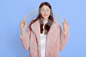 Beautiful woman in pink faux fur coat showing sign ok, being calm, trying to relax, keeping eyes closed, standing isolated over