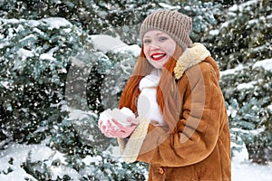 Beautiful woman play with snow on winter outdoor, snowy fir trees in forest, long red hair, wearing a sheepskin coat