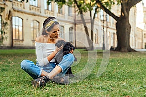 Beautiful woman playing with a black little dog sitting on the grass in the park. Female owner with dog breed toy poodle resting