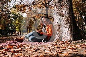 A beautiful woman reading a book in a sunny autumnal day. She is sitting and leaning against a tree while having a book on her