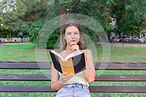 Beautiful woman is reading a book and thinking about something on the bench in the park. Girl has some thoughts and ideas