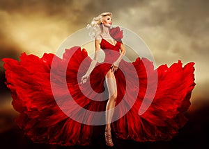Beautiful Woman in Red Dress flying on Wind over Fantasy Sky Background. Fashion Model in Evening Luxury Gown with Slit. Sexy