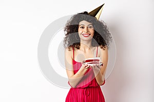 Beautiful woman in red dress, wearing party hat and celebrating birthday, holding b-day cake and making wish, smiling at