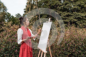 Beautiful woman in red dress white shirt, in summer park forest, draws a picture, white canvas paint and a brush, free