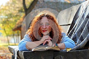 Beautiful woman with red hair is lying on a bench with a book and yellow leaves and looking into the camera. Autumn park backgroun