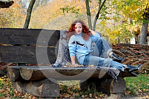 Beautiful woman with red hair sits on a bench and reads a book that lies nearby. Autumn park background. Nearby is a warm blanket