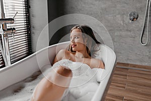 Beautiful woman relaxing in bathtub filled with foam, relaxed lady sitting in hot bath and looking at window