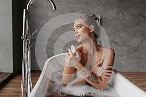 Beautiful woman relaxing in bathtub and looking at window, calm young lady sitting in hot bath in modern bathroom