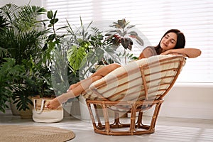 Beautiful woman resting on papasan chair near houseplants indoors. Interior design